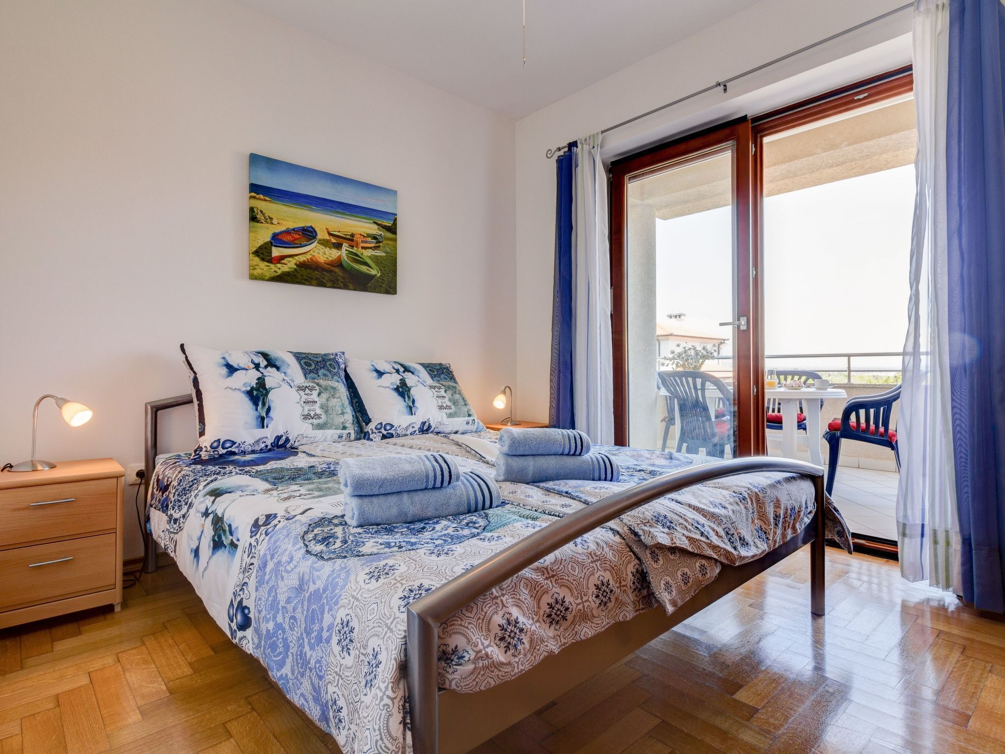 A bedroom with a bed, a nightstand, a lamp, a window with curtains, and a balcony with chairs.