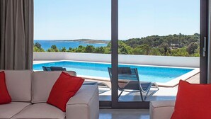 Ein modernes Zimmer mit einer großen Schiebeglastür, die zu einem Poolbereich führt und Meer und Grünlandschaft zeigt.