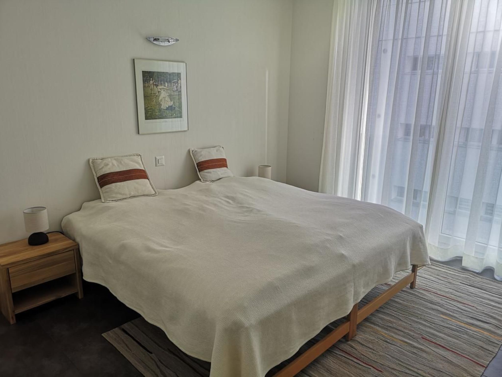 Een opgemaakt bed met een witte dekbedovertrek en twee kussens, een bedlampje, een ingelijst beeld aan de muur en dunne gordijnen voor het raam.