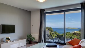 Uma sala de estar moderna com uma TV grande, uma mesa de centro de vidro e um sofá com almofadas laranja. Há uma planta em vaso e um armário branco. O quarto tem uma grande porta de vidro deslizante que dá para uma varanda com vista para o mar.