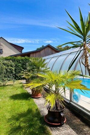 Ein Gewächshaus mit Glasdach, umgeben von Kübelpflanzen und einem Schwimmbecken.