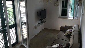 Un salon avec un canapé, un téléviseur, un lustre et une fenêtre avec des volets.