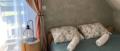 Une chambre confortable avec un lit, des oreillers, une petite table d’appoint en bois, un tapis moelleux et une fenêtre avec des rideaux.