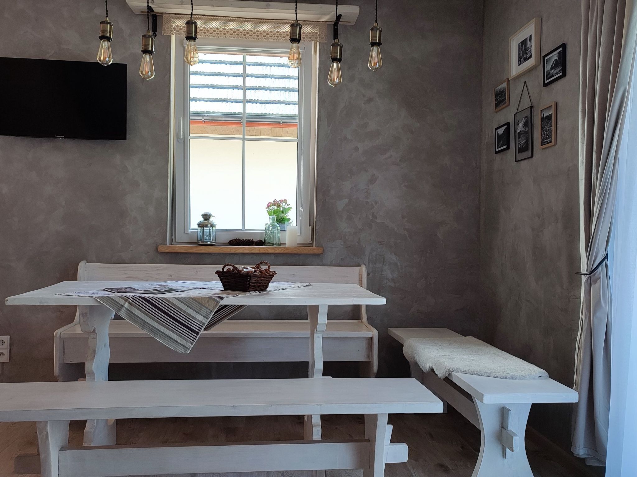 Een eetruimte met een witte tafel en bank, een raam met zonwering, hangende lampen en ingelijste foto's aan de muur.
