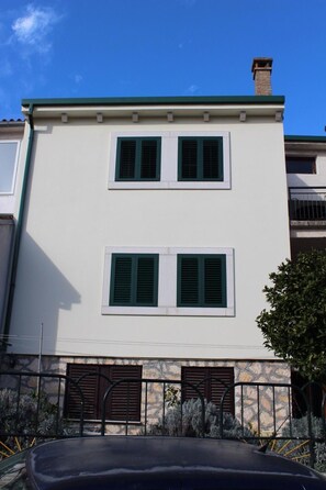 Un edificio de dos pisos con techo plano y chimenea. Cada planta tiene dos ventanas con contraventanas verdes.