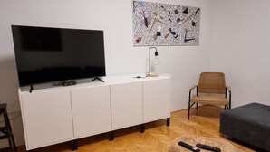 Un salon moderne doté d’un téléviseur à écran plat, de meubles blancs, d’un pouf en fibres tressées, d’un fauteuil et d’un parquet en bois à motifs chevrons.
