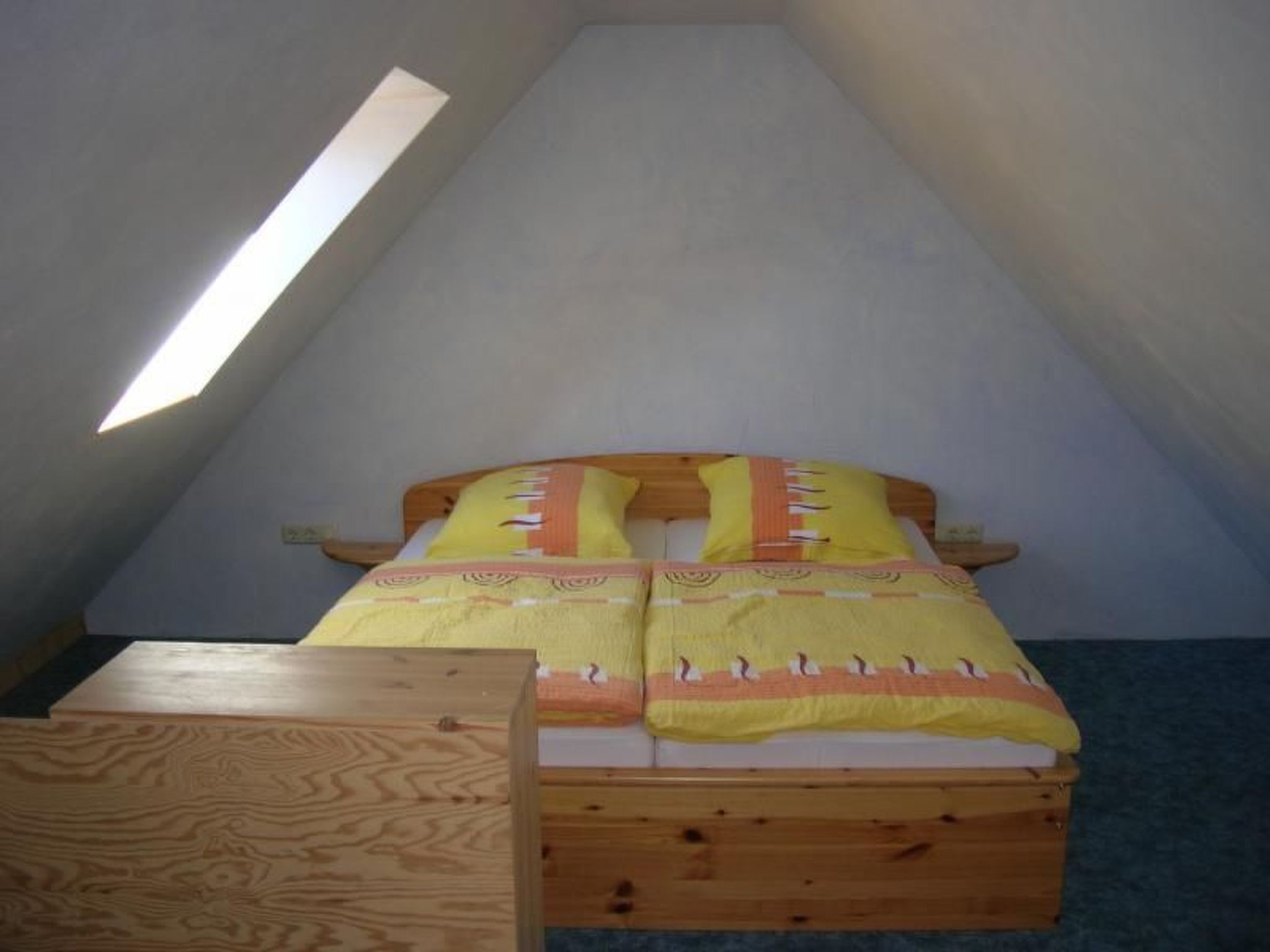 Ein kleines, gemütliches Schlafzimmer im Dachgeschoss mit schrägen Wänden, einem hölzernen Bett mit gelber Bettwäsche und einem kleinen Holztisch.