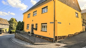 A yellow building with a corner entrance, a visible street sign, and a paved area with a metal railing. - Siebers by Interhome (Altendorf)