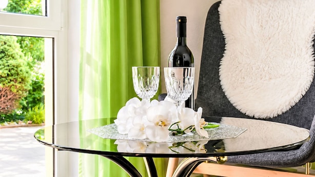 A glass table with a bottle and two glasses, a white orchid, and a chair with a white fur throw. - Siebers by Interhome (Altendorf)