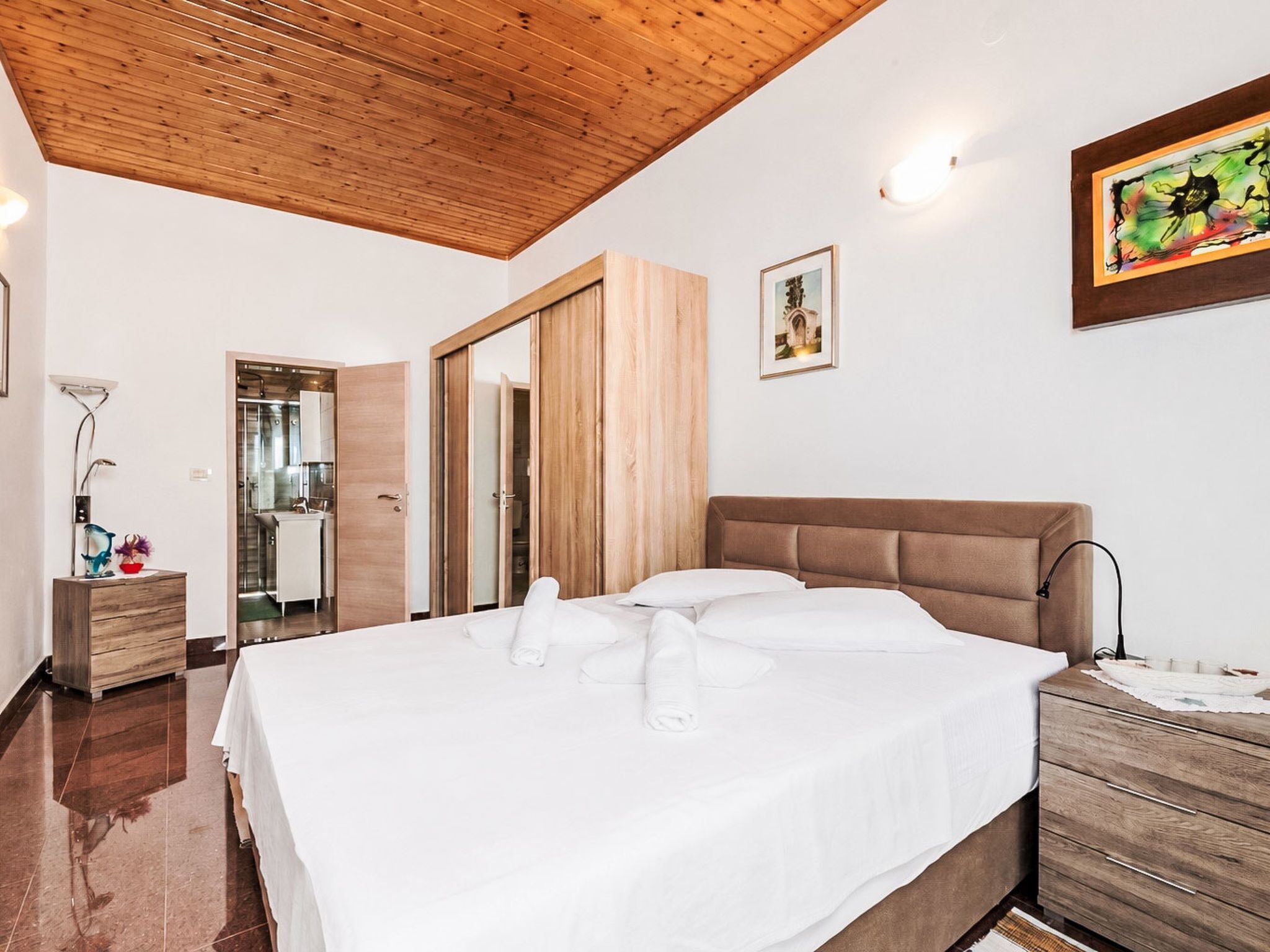 A bedroom with a wooden ceiling, a bed with white linens, a wooden dresser, and a painting on the wall.