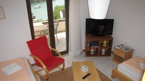Une chambre d’hôtel avec un fauteuil rouge, une table en bois, un téléviseur, une bibliothèque et une vue sur la mer, visible à travers la porte-fenêtre coulissante.