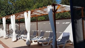 Zona de piscina con tumbonas bajo una pérgola con cortinas blancas.