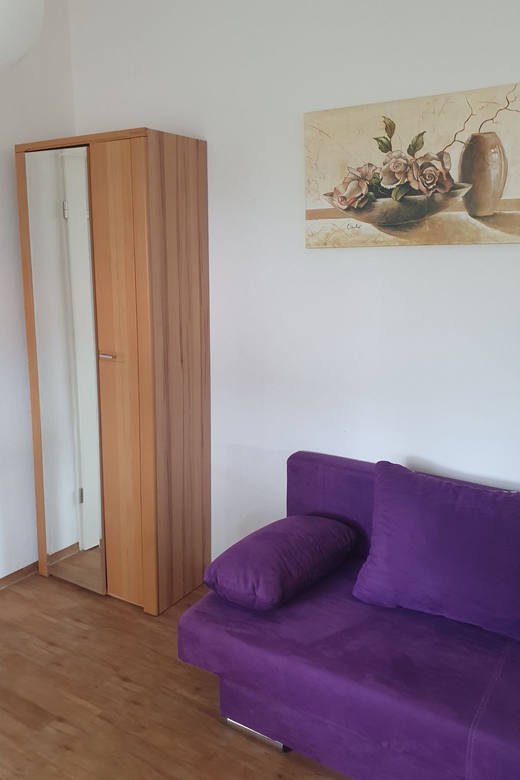 A room with a purple sofa, a wooden wardrobe, and a painting of flowers on the wall.