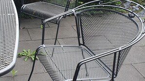 Outdoor metal chairs and a table on a paved patio with a garden in the background.