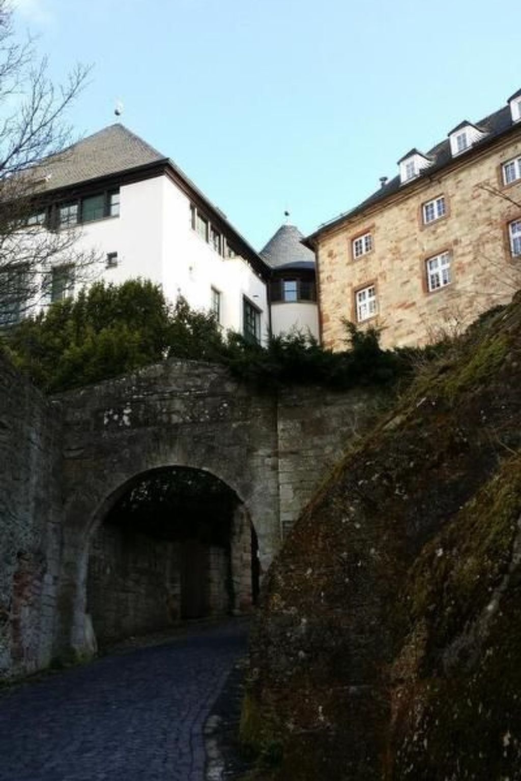 A stone archway leading to a building with a white facade and a stone building with a pitched roof.