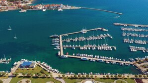 Oversigtsbillede af en lysthavn med mange både i havnen, omgivet af en kystby med synlige bygninger og en klar, blå himmel.