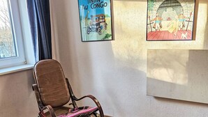 Una habitación con piso de madera, una silla marrón con un cojín colorido, una ventana con cortinas y dos cuadros en la pared.