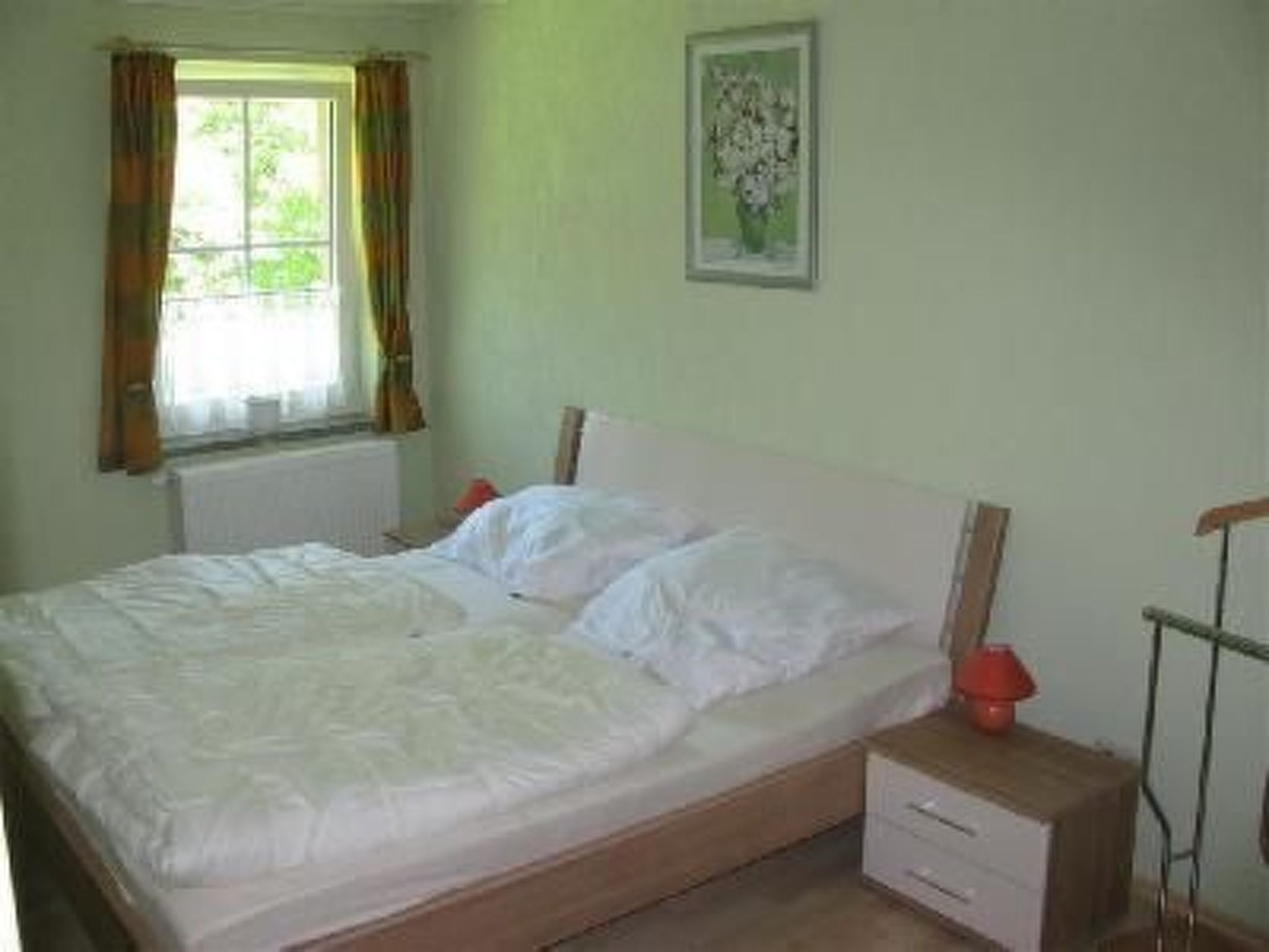 Une chambre à coucher avec un lit, une table de chevet, une fenêtre avec des rideaux et un tableau encadré au mur.