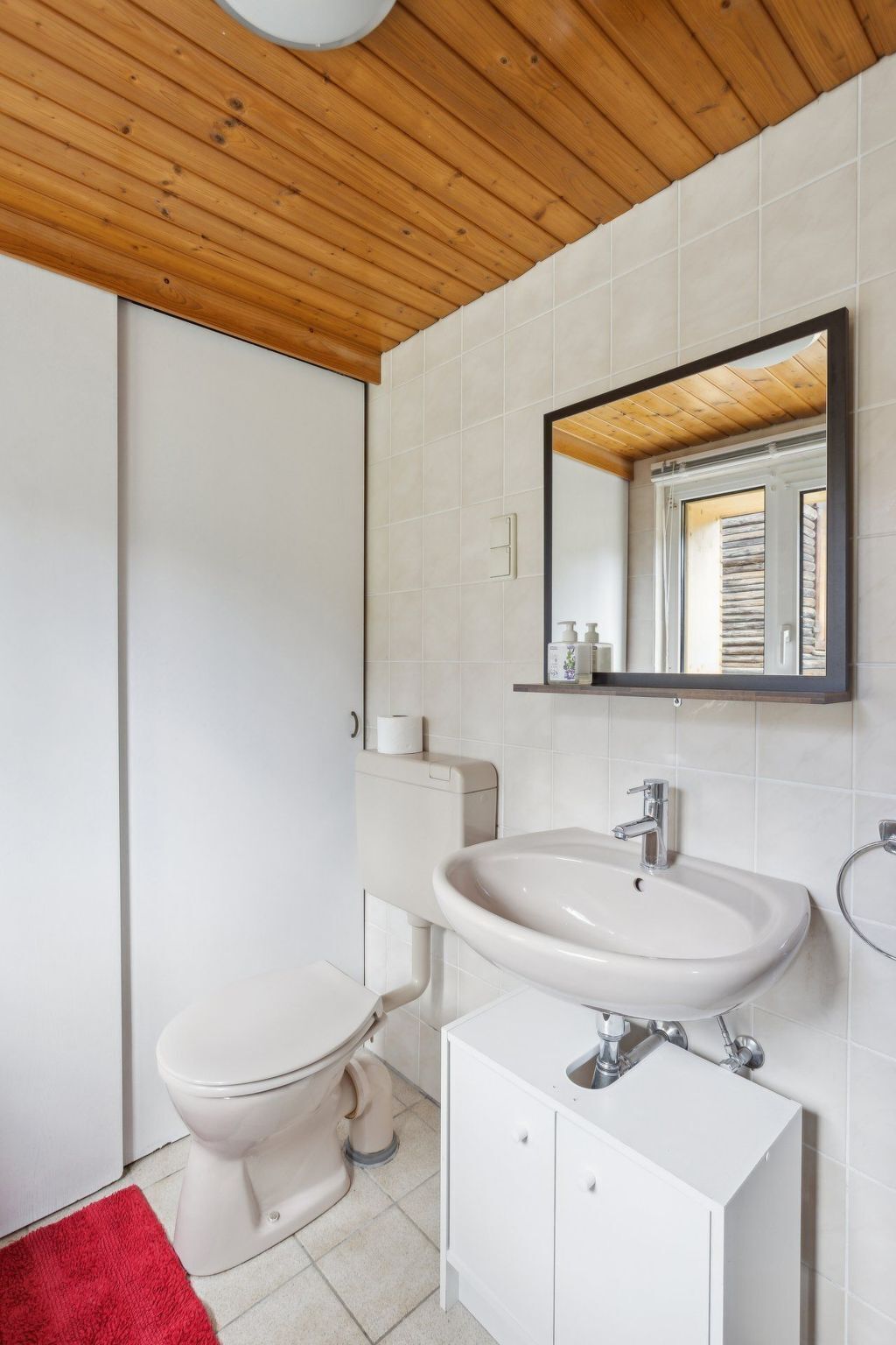 Ein Badezimmer mit einer weißen Toilette, einem Waschbecken mit Spiegel darüber und einer roten Fußmatte auf dem Boden.