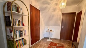 Um corredor com uma estante de livros, uma mesinha com objetos decorativos e duas portas de madeira.