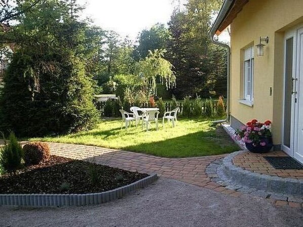 A patio with a brick pathway leading to a white table and chairs, surrounded by lush greenery and a yellow building with a door and windows. - J. v. Eichendorff by Interhome (Born)