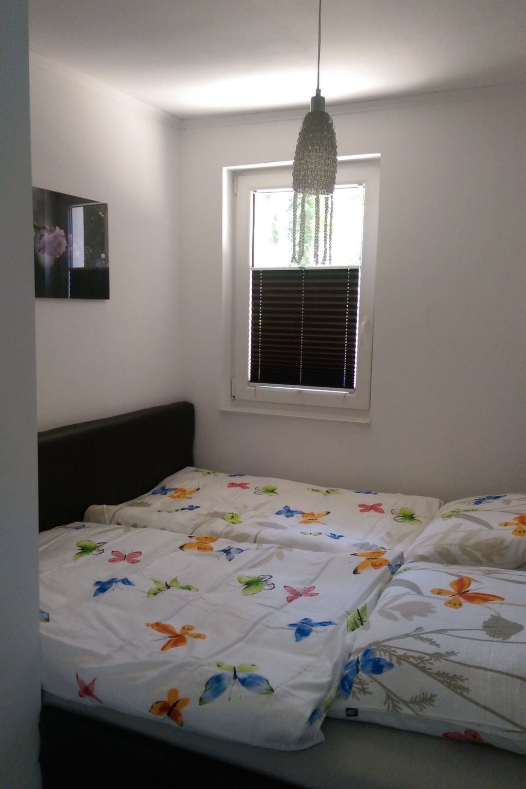 A bedroom with a bed, a window with blinds, a hanging light fixture, and a framed picture on the wall.