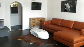 A modern living room with a leather sofa, a white pouf, a wooden cabinet, and a large painting on the wall.