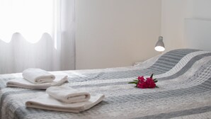 Un lit bien fait, avec des serviettes pliées et une fleur décorative, une lampe de chevet et une fenêtre ornée de rideaux légers.