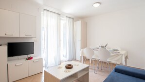Une pièce moderne et bien éclairée, dotée d’un téléviseur à écran plat, d’une table à manger avec des chaises, d’un canapé et d’une fenêtre ornée de rideaux légers.