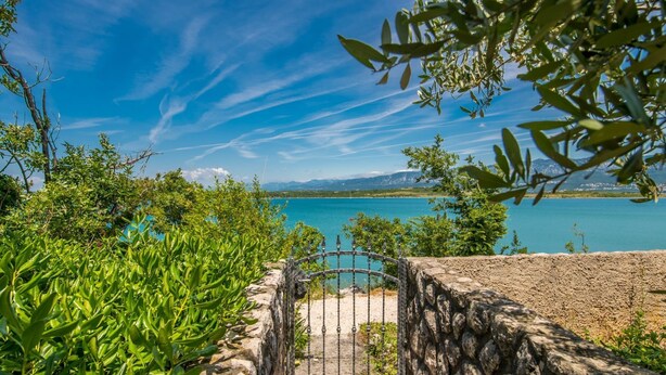 A stone pathway leading to a metal gate, surrounded by lush greenery and overlooking a calm lake with mountains in the distance. - Biserka by Interhome (Krk/Čižići)