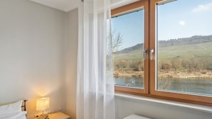 A bedroom with a bed, a nightstand with a lamp, a window with curtains, and a view of a river and hills. - mit Balkon und schönem Moselblick by Interhome (Klüsserath)