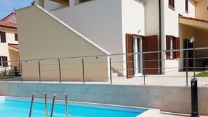 Ein Schwimmbecken mit einer Metallleiter, einer gefliesten Terrasse und einem Gebäude mit einer beige farbenen Fassade und Holzläden.