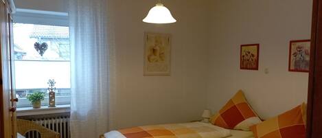 A bedroom with two beds, a window with a view of buildings, a radiator, and a hanging light fixture.