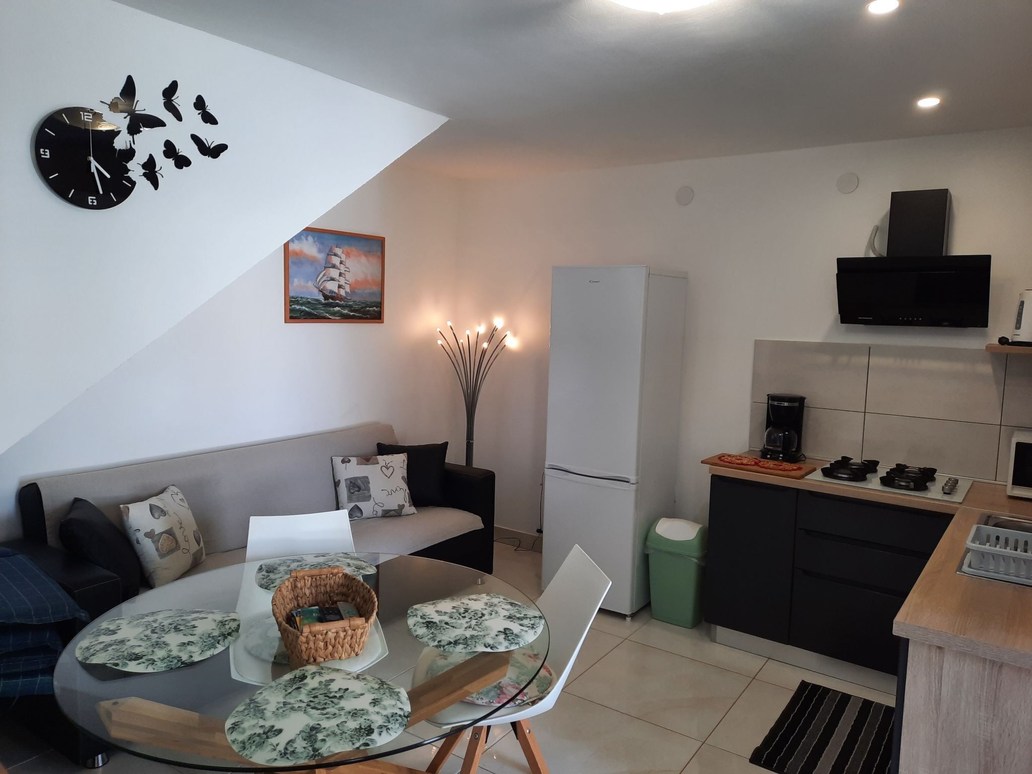 Une cuisine moderne équipée d’un réfrigérateur, d’une cuisinière et d’un four à micro-ondes. On y trouve une table à manger avec des chaises, ainsi qu’une table basse en verre. Un canapé et une horloge murale décorative sont également visibles.