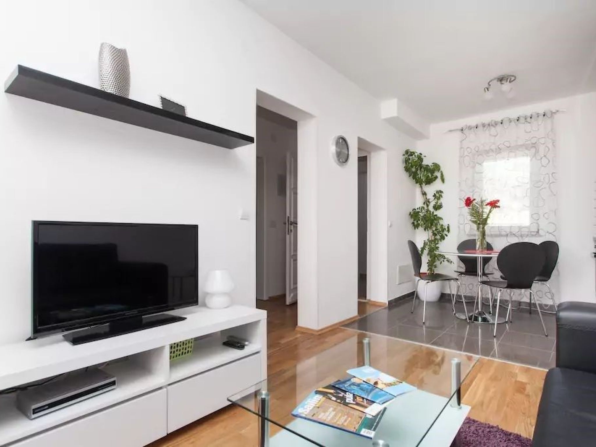 Uma sala de estar moderna com televisão de tela plana, móvel de entretenimento branco, mesa de centro de vidro e uma área de jantar com mesa e cadeiras.