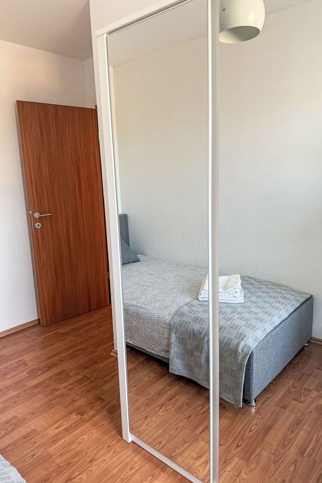 A bedroom with a wooden door, a bed with a grey and white patterned bedspread, and a white ceiling light.