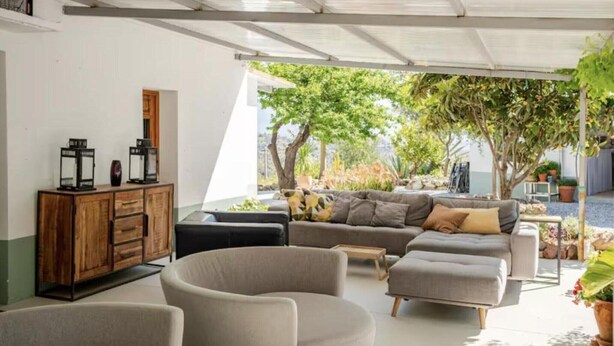 A modern living room with a sofa, ottoman, and chairs, a wooden cabinet, and a view of a garden with trees and plants. - 5-Bedroom Villa with Pool, Swimjet and Jacuzzi (Comares)