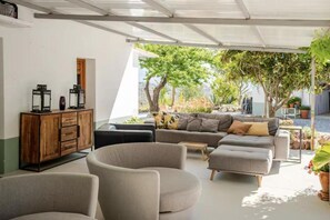 A modern living room with a sofa, ottoman, and chairs, a wooden cabinet, and a view of a garden with trees and plants. - 5-Bedroom Villa with Pool, Swimjet and Jacuzzi (Comares)