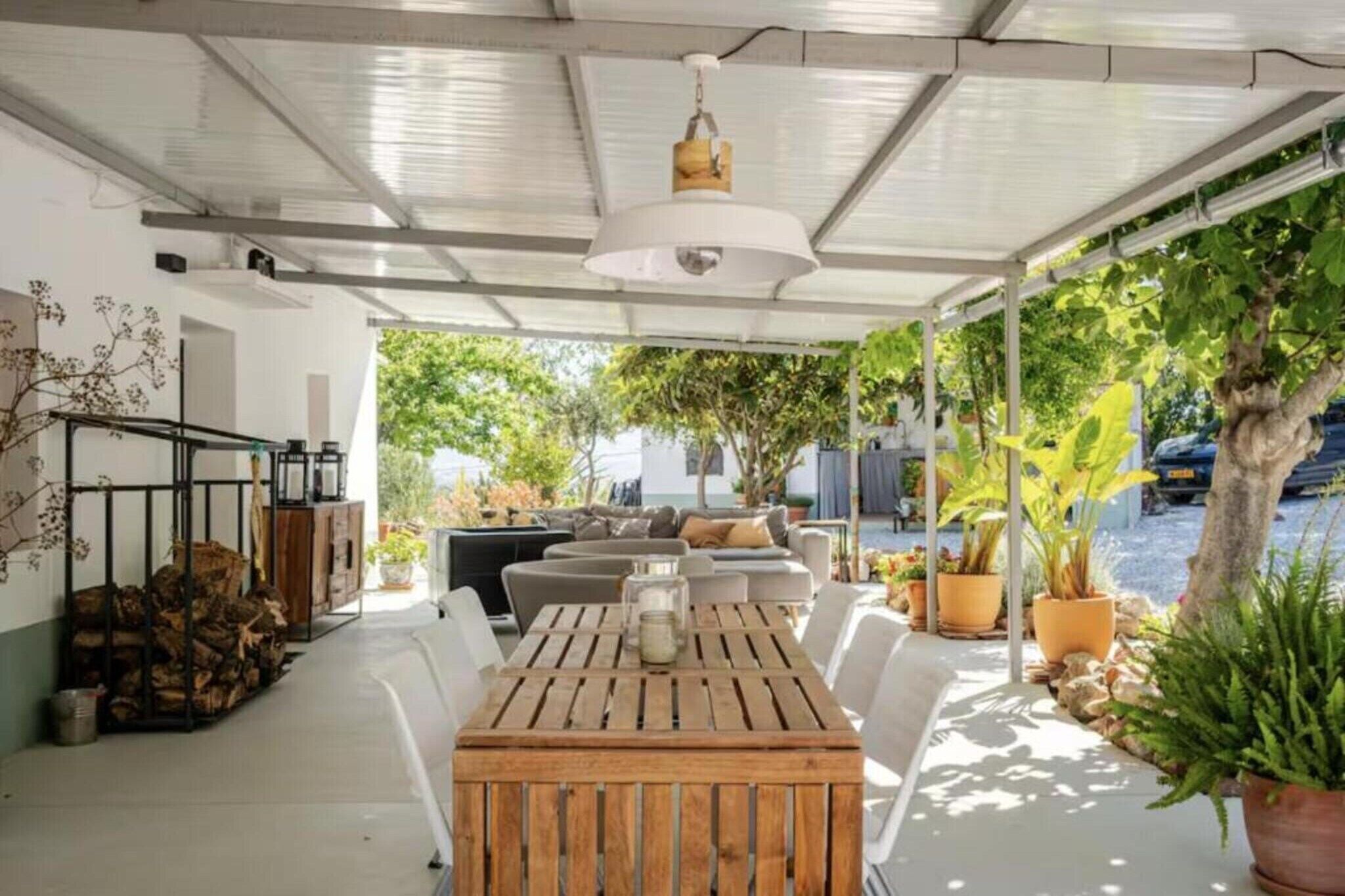 Un patio al aire libre con una mesa y sillas de madera, rodeado de plantas en macetas y un sofá.
