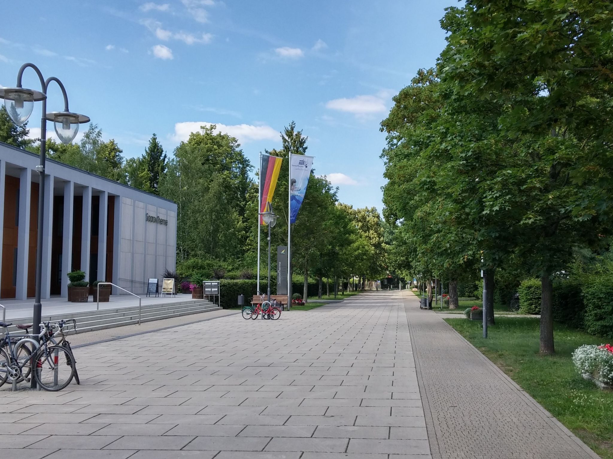 Een verharde looppad met een modern gebouw aan de linkerkant, een vlaggenmast met vlaggen, en fietsen geparkeerd aan de linkerzijde.