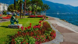 A park with a statue, benches, and a flower bed by a waterfront. - Haus Antonini by Interhome (Brissago)