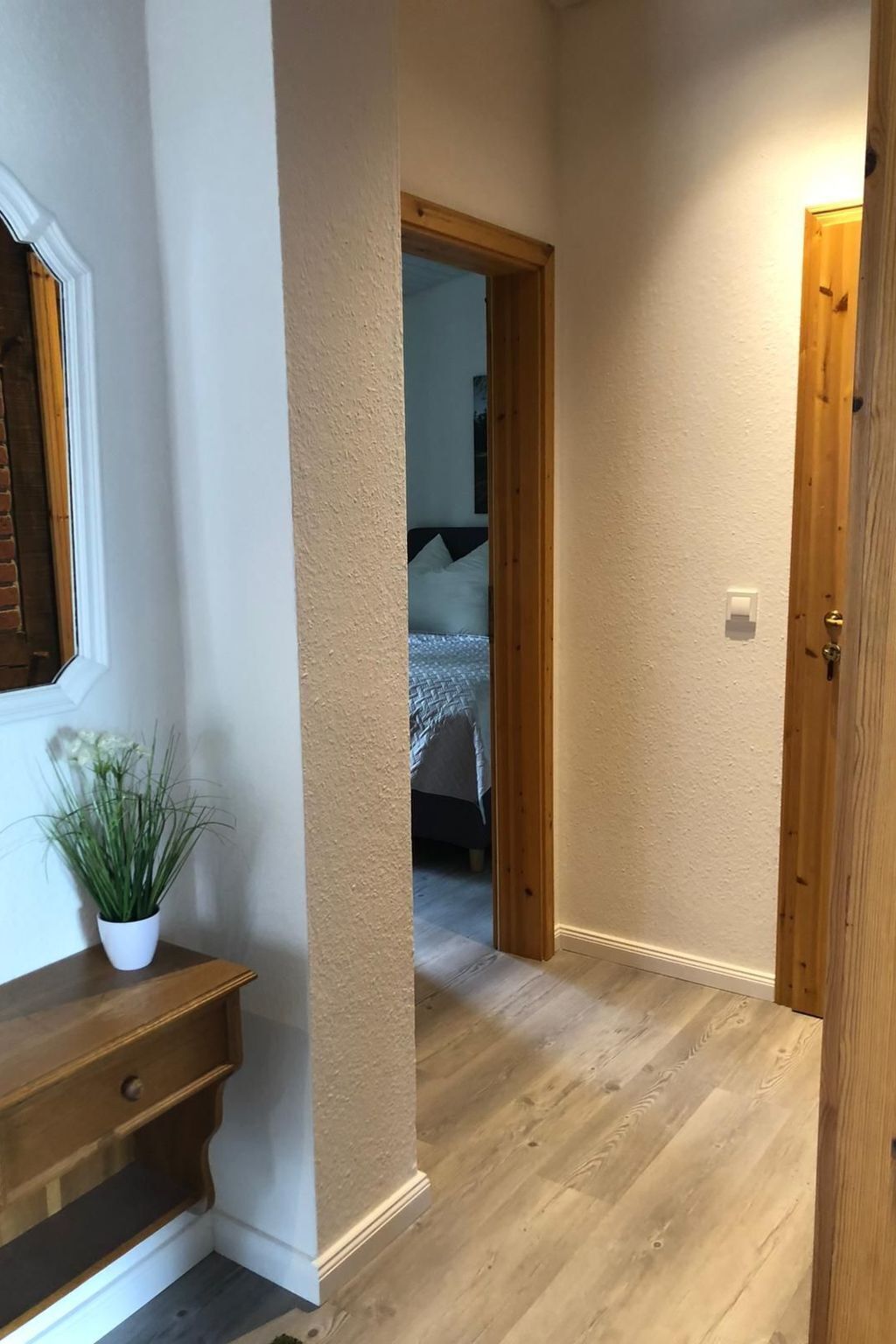 En hall med en trätrappa, en krukväxt och en säng som syns genom en öppen dörr.