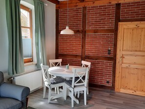 Ein gemütlicher Raum mit Backsteinwand, einer Holztür, einem runden Tisch mit weißen Stühlen und einer Pendelleuchte.