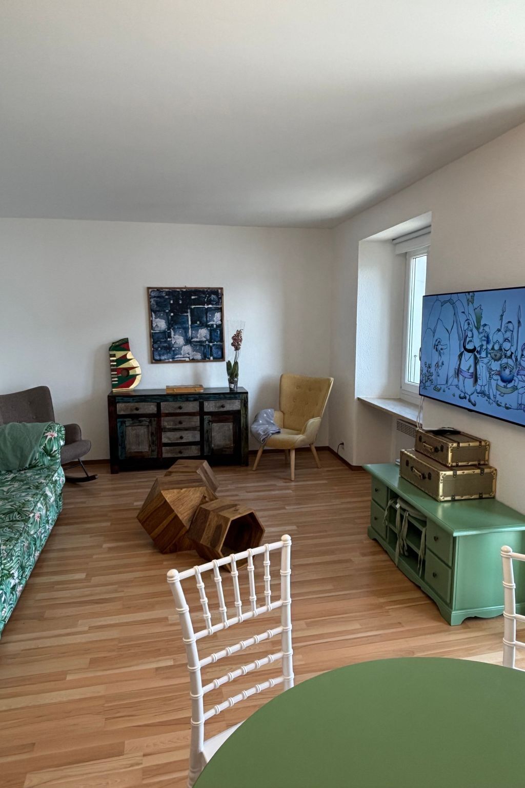 A living room with a sofa, a wooden coffee table, a green table, a white chair, a cabinet, and a TV on the wall.