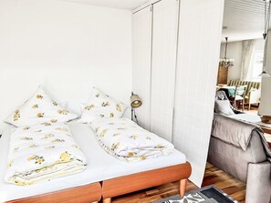 A bedroom with a wooden bed, white bedding with floral patterns, a bedside lamp, and a view into a dining area. - Wohnung in Bergen und Panoramablick by Interhome (Bergen)