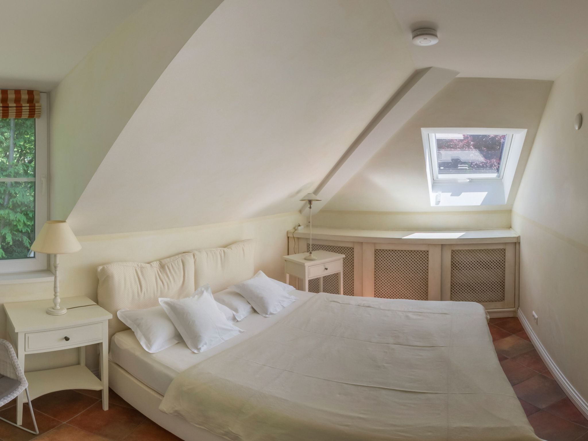 Un dormitorio con techo inclinado, una cama con ropa blanca, un escritorio con lámpara y una ventana con vistas a árboles.