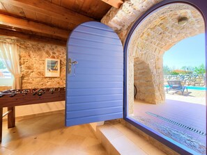 Une pièce avec un mur en pierre, un plafond en bois, une porte bleue et une vue sur un espace extérieur avec piscine.