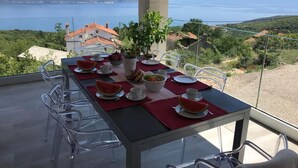 A dining table with chairs, plates, cups, and a bowl of fruit, set on a balcony with a view of a body of water and hills. - Vacation home Stepča by Interhome (Brseč)