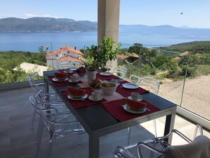 A dining table with chairs, plates, cups, and a bowl of fruit, set on a balcony with a view of a body of water and hills. - Vacation home Stepča by Interhome (Brseč)