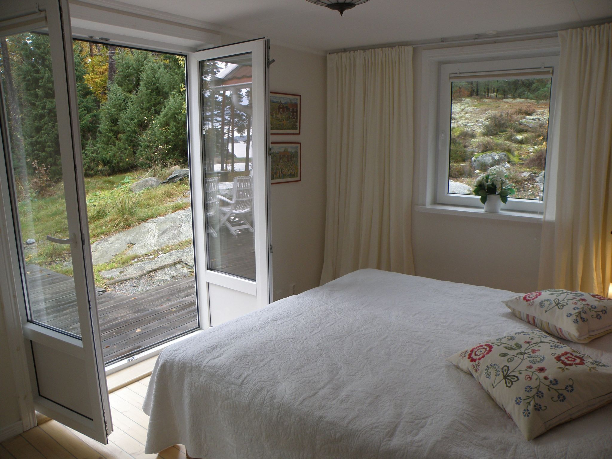 Une chambre avec un lit, une fenêtre avec des rideaux, une porte donnant sur un espace extérieur et une petite plante posée sur le rebord de la fenêtre.